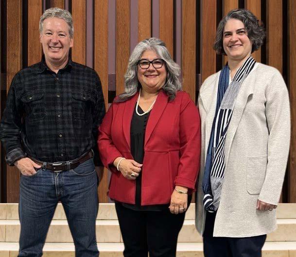 Three people standing together in a group photo: a person in plaid shirt and jeans, a person in red blazer and black pants, and a person in cream-colored sweater and black pants.