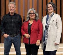 Three people standing together in a group photo: a person in plaid shirt and jeans, a person in red blazer and black pants, and a person in cream-colored sweater and black pants.